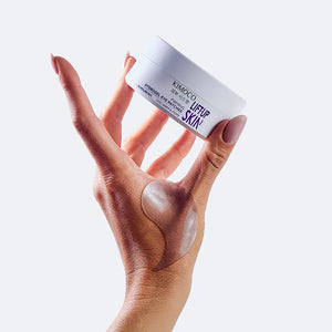 Uniesiona dłoń trzymająca pojemnik "Kimoco Lift-Up Skin Firming Hydrogel Eye Patches" między palcami, z płatkiem pod oczy nałożonym na wierzchu dłoni, na jasnym tle.