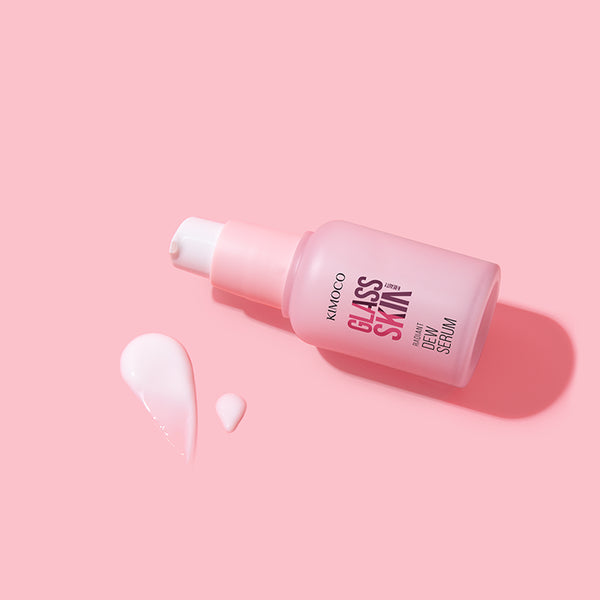 Różowa butelka z pompką kosmetyku Kimoco Glass Skin Radiant Dew Serum leżąca ukośnie, z lewej rozsmarowana kropla i mniejsza kropelka produktu, na różowym tle.
