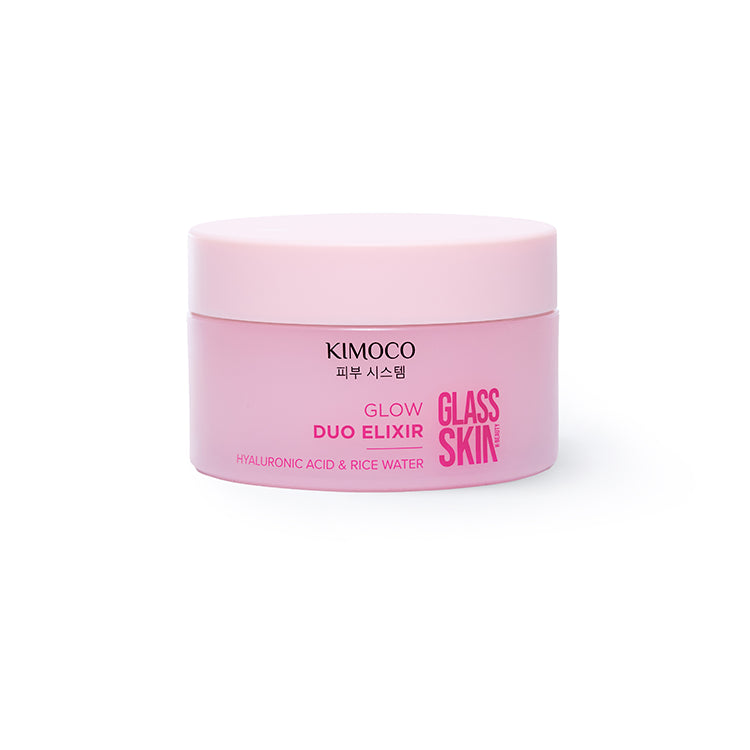 Zdjęcie na jasnoszarym tle: u góry dłoń dociskająca stos produktów "Kimoco Glass Skin Glow Duo Elixir" złożony z dwóch słoiczków i pudełka pomiędzy nimi, u dołu druga dłoń podtrzymująca stos od spodu.