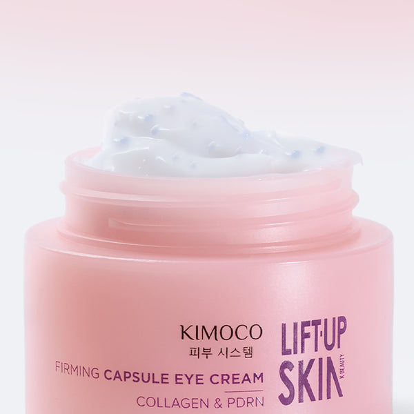 Otwarty słoiczek "Kimoco Lift-Up Skin Firming Capsule Eye Cream" z widocznym białym kremem z drobnymi niebieskimi kapsułkami, na jasnoróżowym tle