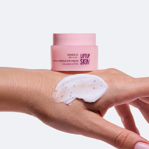 Słoiczek "Kimoco Lift-Up Skin Firming Capsule Eye Cream" stojący na wierzchu dłoni z rozsmarowanym białym kremem z drobnymi kapsułkami, na jasnoszarym tle.