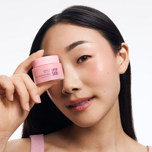 Kobieta o azjatyckich rysach twarzy i ciemnych włosach, trzymająca z lewej strony przy oku różowy słoiczek kosmetyku Kimoco Lift-Up Skin Firming Capsule Eye Cream, na białym tle.