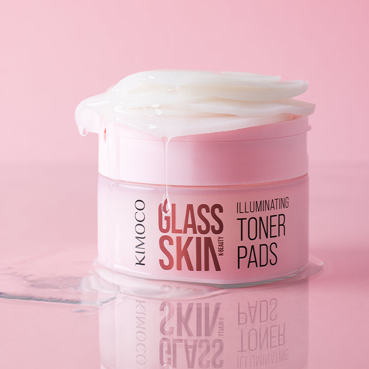 Pojemnik "Kimoco Glass Skin Illuminating Toner Pads" z płatkami wystającymi z wieczka i ściekającym płynem, z odbiciem na lustrzanej powierzchni, na różowym tle.