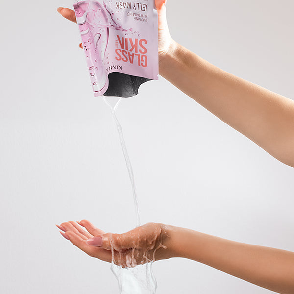 U góry dłoń trzymająca odwrócone opakowanie "Kimoco Glass Skin Jelly Mask" z wypływającym płynem, u dołu druga dłoń łapiąca ściekający płyn, na jasnoszarym tle.