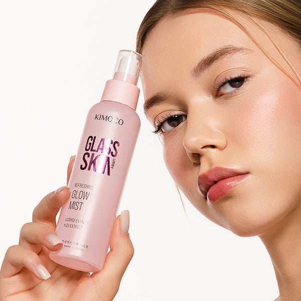 Twarz kobiety z jasną, promienną cerą, trzymająca przy policzku różową butelkę z rozpylaczem kosmetyku Kimoco Glass Skin Refreshing Glowy Mist, na białym tle.