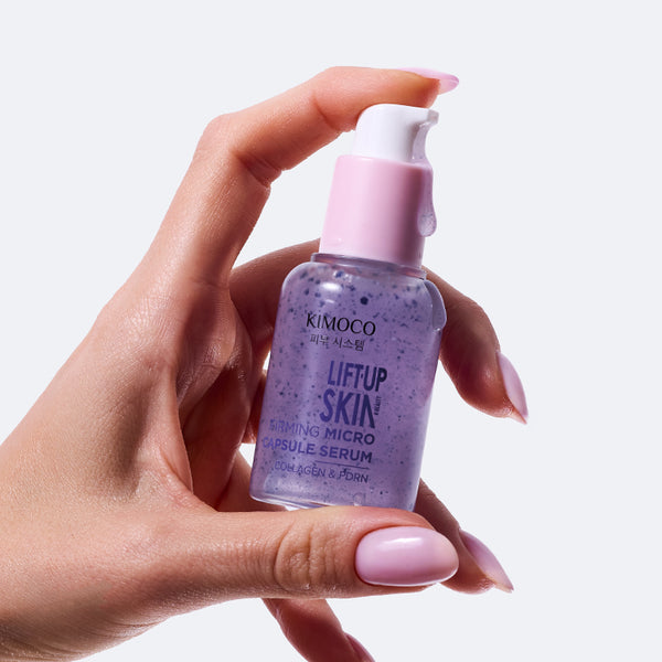 Dłoń trzymająca butelkę "Kimoco Lift-Up Skin Firming Micro Capsule Serum" z fioletowym serum, z kroplą płynu na dozowniku, na jasnoszarym tle.