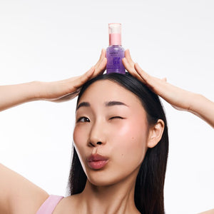 Kobieta o azjatyckich rysach twarzy i ciemnych włosach, mrugająca okiem, trzymająca oburącz nad głową fioletową butelkę kosmetyku Kimoco Lift-Up Skin Firming Micro Capsule Serum.