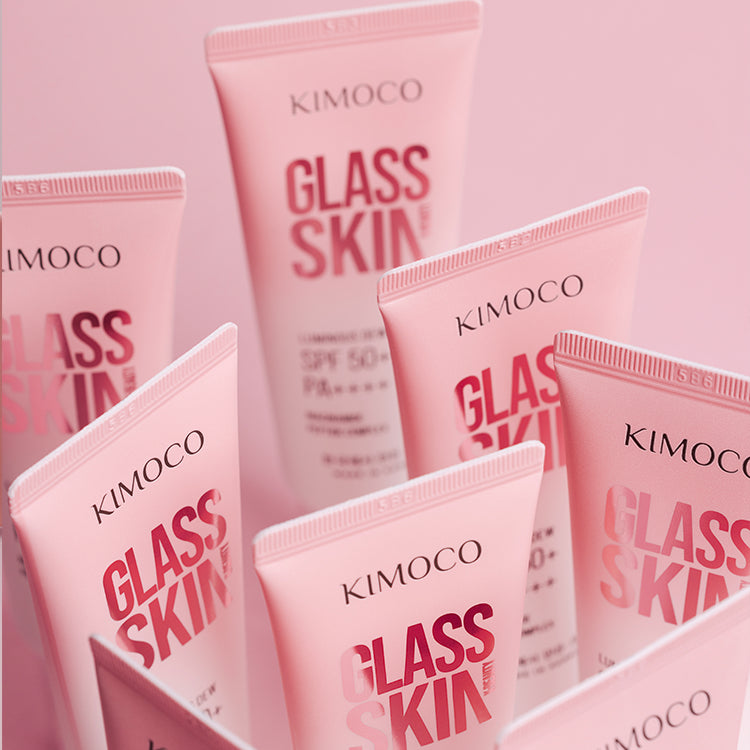 Zdjęcie na białym tle: u góry twarz kobiety z rumieńcem na policzku i dłonią z kremem na palcach uniesioną przy twarzy, u dołu tuba "Kimoco Glass Skin Luminous Dew SPF 50+ PA+++" trzymana w drugiej dłoni.