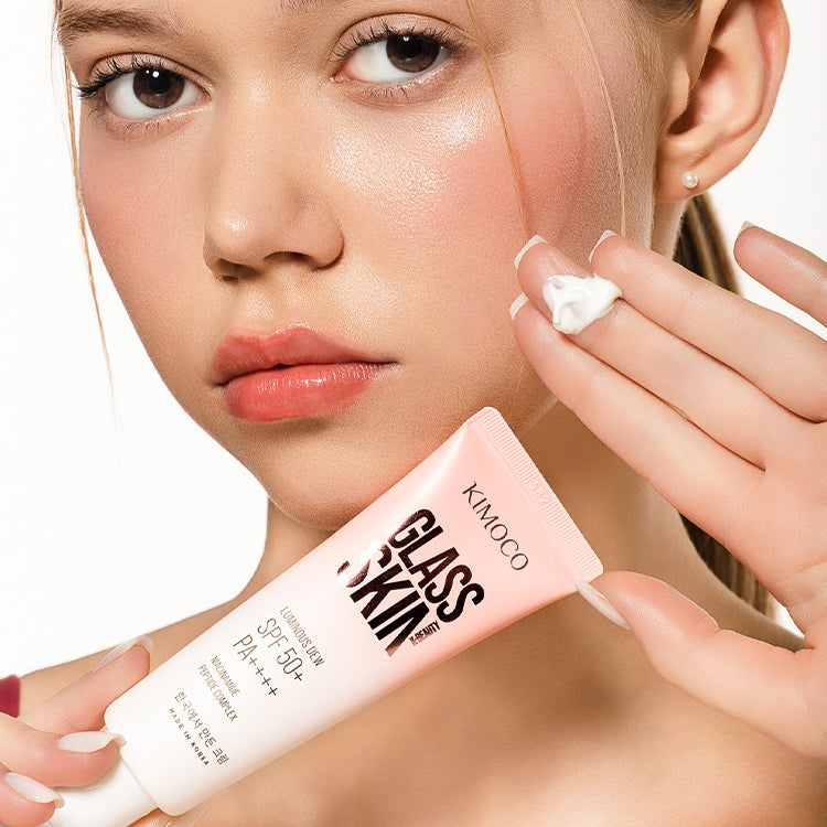 Zdjęcie na białym tle: u góry twarz kobiety z rumieńcem na policzku i dłonią z kremem na palcach uniesioną przy twarzy, u dołu tuba "Kimoco Glass Skin Luminous Dew SPF 50+ PA+++" trzymana w drugiej dłoni.