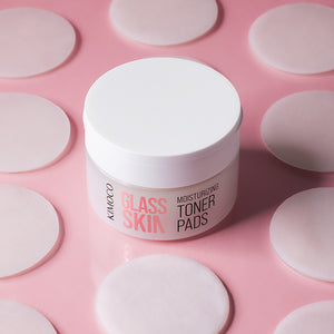 Zamknięty pojemnik "Kimoco Glass Skin Moisturizing Toner Pads" otoczony rozłożonymi okrągłymi płatkami, na różowym tle.
