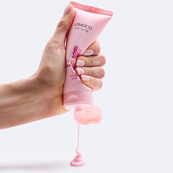 Dłoń trzymająca różową tubkę kosmetyku Kimoco Glass Skin Glow & Lift Wrapping Mask, z otworu wypływa różowa konsystencja produktu skapująca w dół, na białym tle.