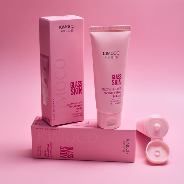 Z lewej stojące pudełko Kimoco Glass Skin Glow & Lift Wrapping Mask, na środku różowa tubka tego samego produktu, u dołu leżące pudełko tej samej marki, z prawej otwarta tubka z widoczną zakrętką, na różowym tle
